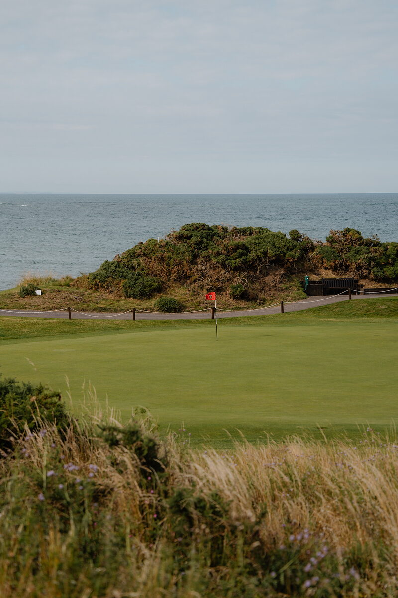 A green marked by a red flag bordered by gorse, the sea stretching to the horizon