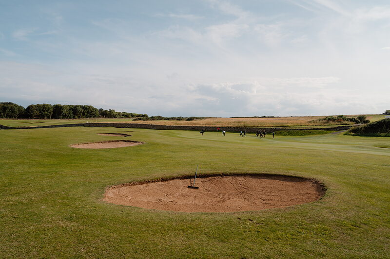 Golfers walking a fairway bordered by bunkers, woodland lining the horizon