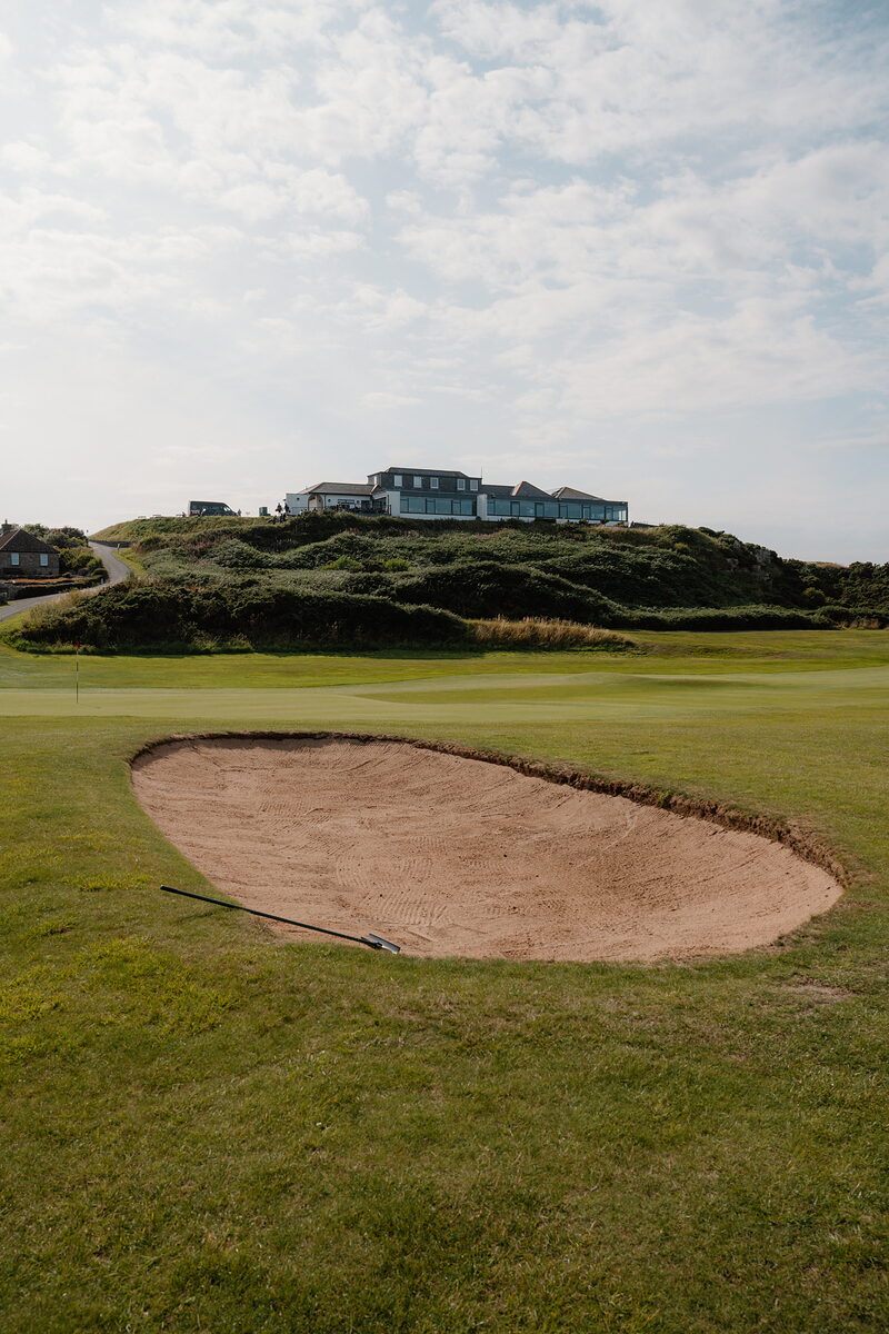 A bunker beside a green, a clubhouse perched on a rise above the dunes with figures gathered on its terrace