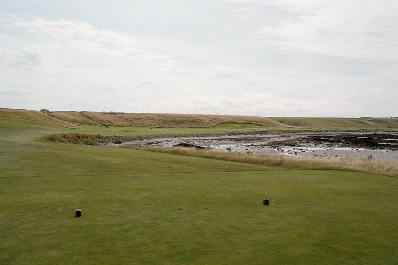 A tee marked by two black markers overlooking a muddy tidal creek bordered by long grass