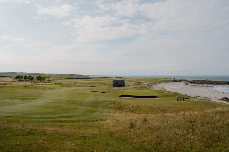 A stone shelter beside the green at Crail, with the sea and a sandy shoreline beyond