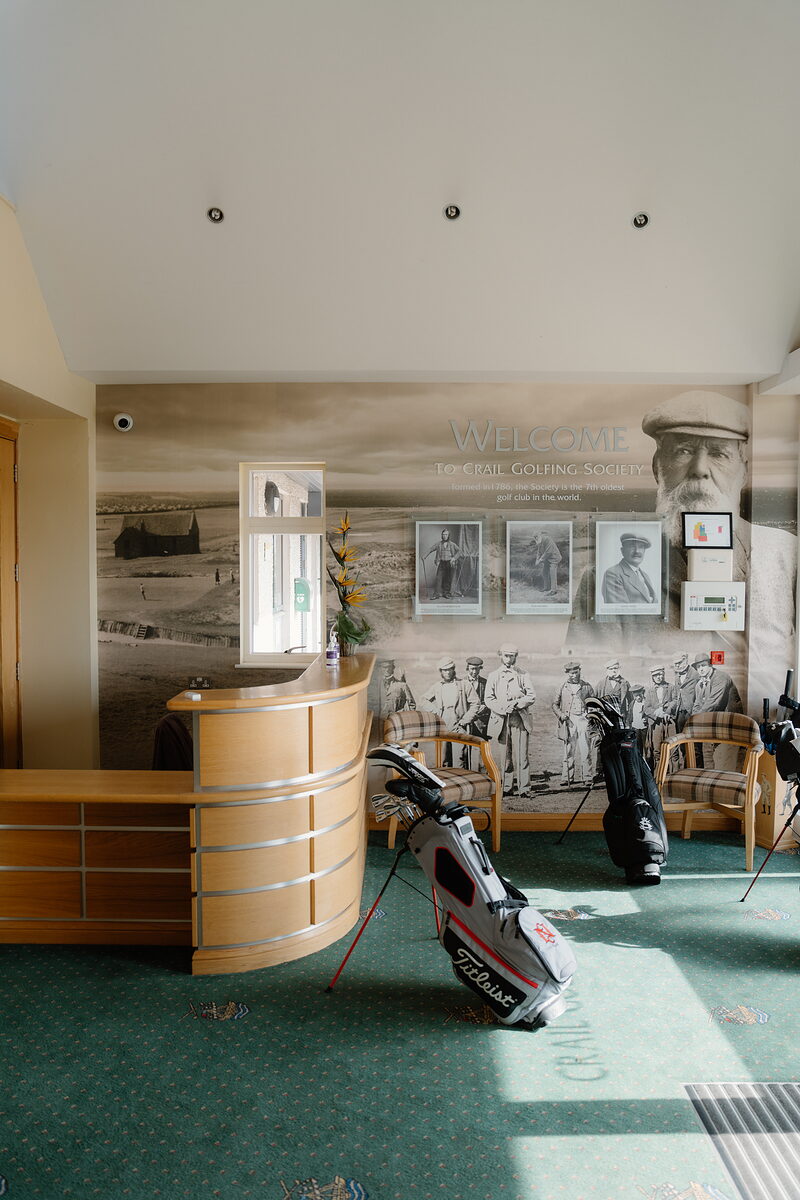 A reception desk beneath a mural welcoming visitors to Crail Golfing Society, golf bags resting against the wall