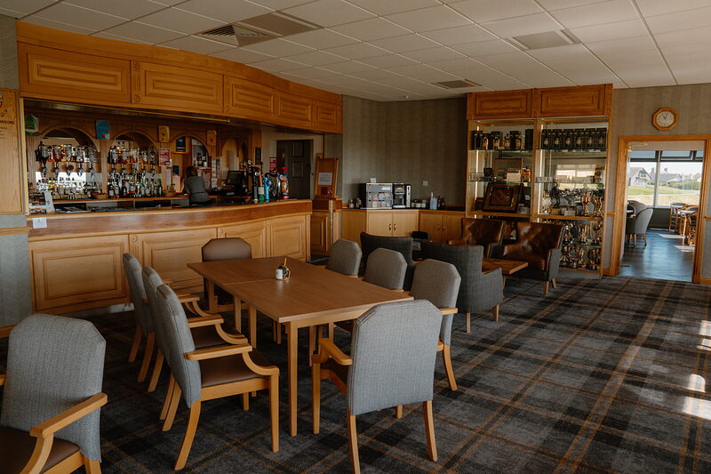 A golf clubhouse bar backed by wooden cabinetry, dining tables and chairs set before it beneath a window view