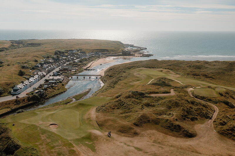 A coastal village and river mouth bordering dune fairways, a footbridge crossing the water