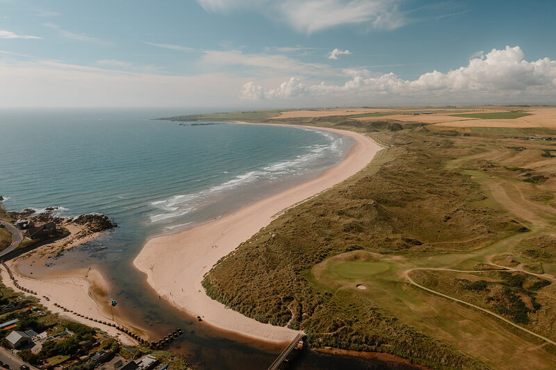 Dune fairways bordering a long sandy beach, a river mouth and harbour visible in the distance