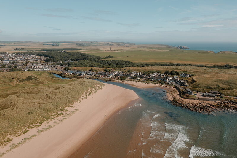 A coastal village and river mouth bordering a sandy beach, dune links stretching along the shore