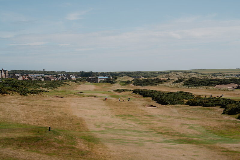 A fairway leading towards dune links and a village, golfers scattered across the course