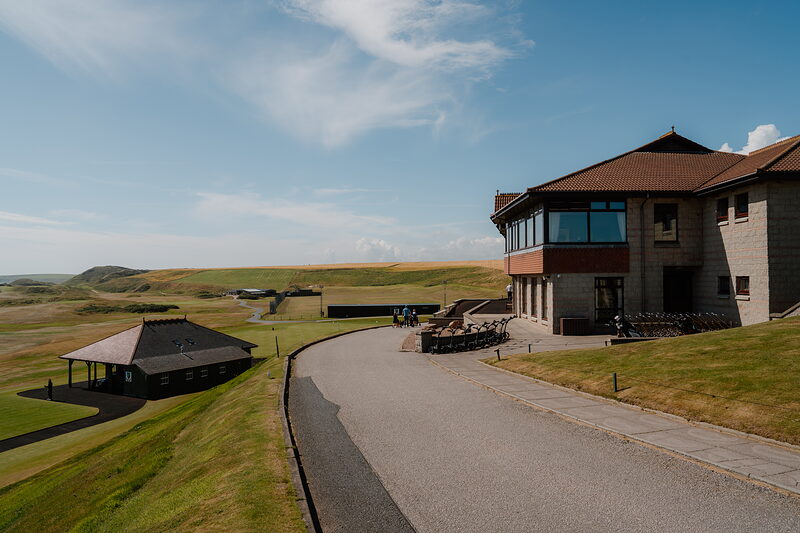 A clubhouse driveway lined with golf trolleys, thatched outbuildings visible across the fairway