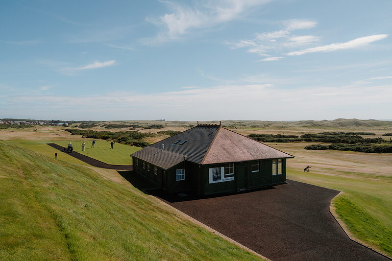 A pitched-roof clubhouse set among dunes, golfers walking the fairway beyond it