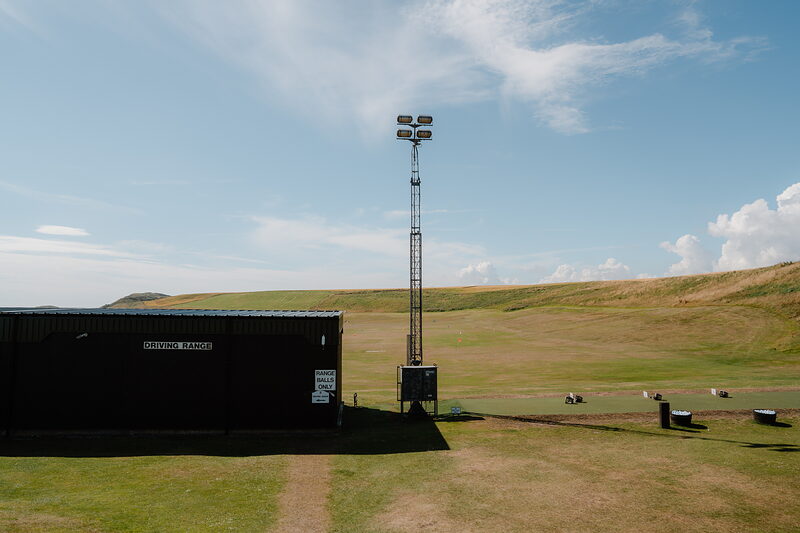 A floodlight tower standing beside a driving range building, sheep grazing on the grass nearby
