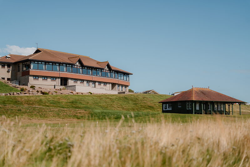 A large timber clubhouse standing on a rise above a smaller pavilion, dune grass in the foreground