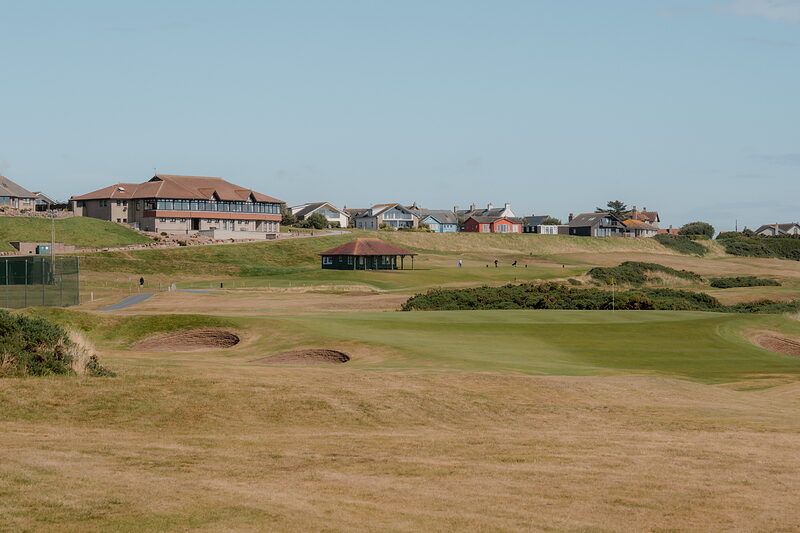 A fairway leading towards a clubhouse and pavilion set among rooftops on the horizon