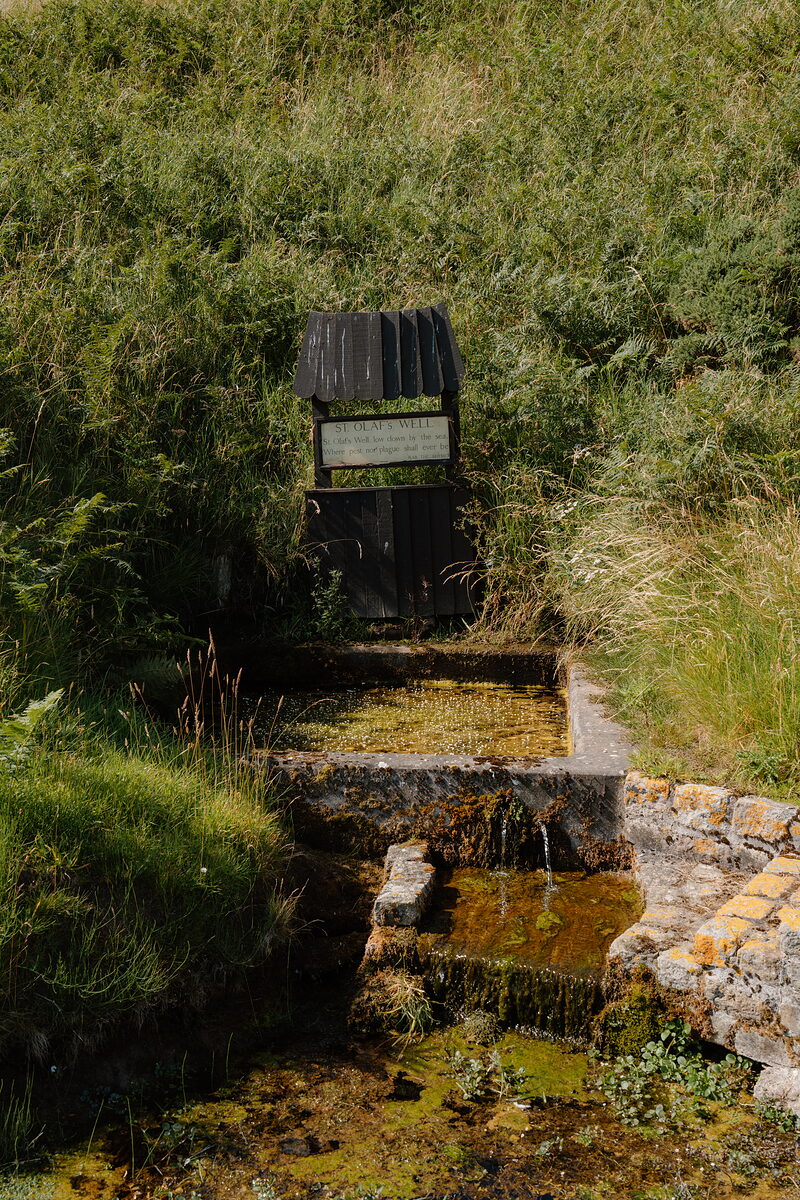 A timber well-house marked St Olaf's Well set into a bank, water pooling in a stone trough below