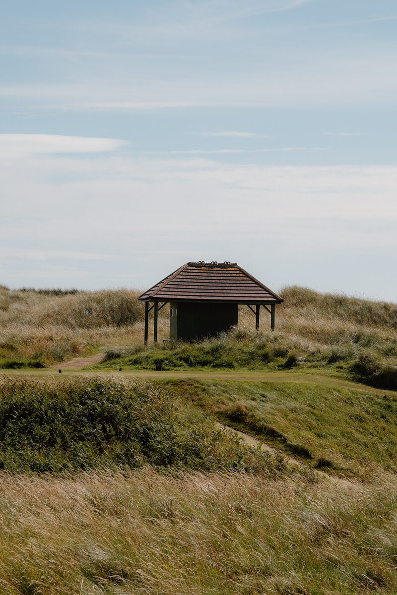 A thatched-roof shelter standing on a mound between fairways, dune grass surrounding it