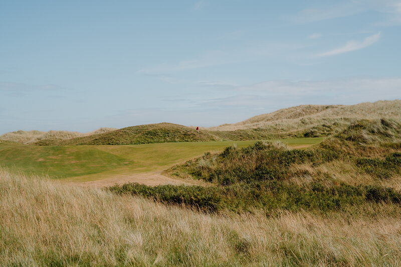 A red flag marking a green set among dunes, dense scrub bordering the fairway