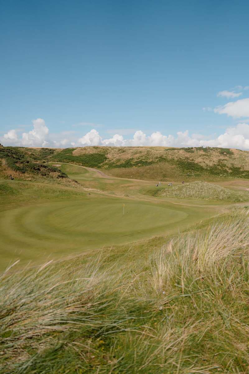 A green set among dunes, distant figures crossing a path in the background