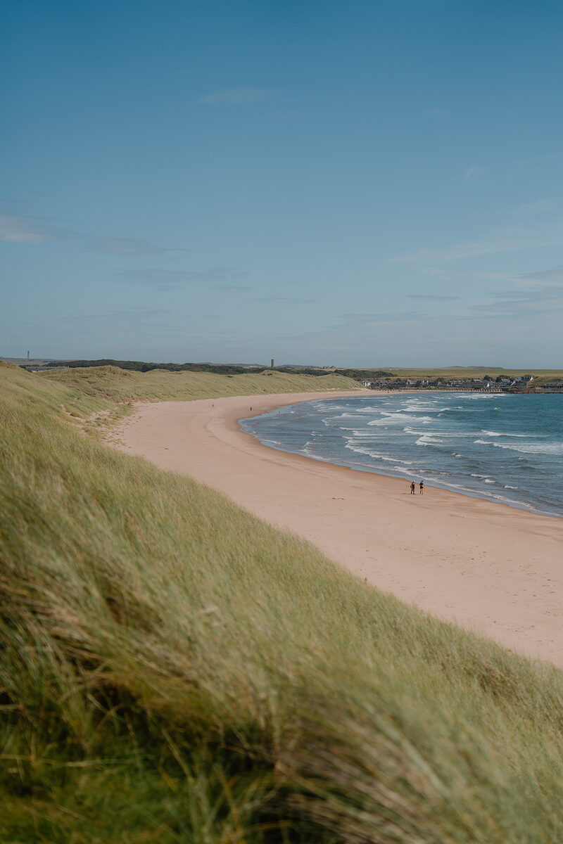 A curving sandy beach beneath grassy dunes, two figures walking near the water's edge