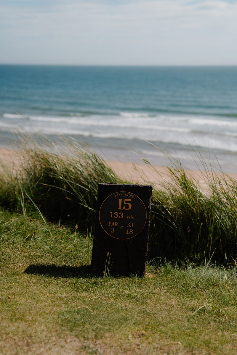 A course marker board for hole 15, 133 yards, standing before a sandy beach and the sea