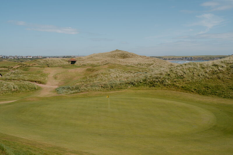 A dark timber shelter set into dunes beside a green, water visible beyond the grassy mounds