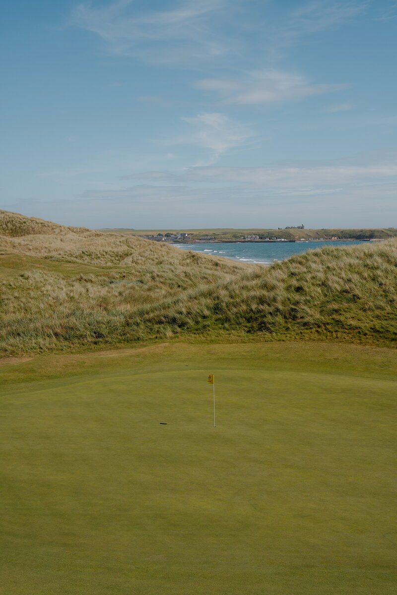 A yellow flag marking a green, a coastal village visible across the bay beyond the dunes