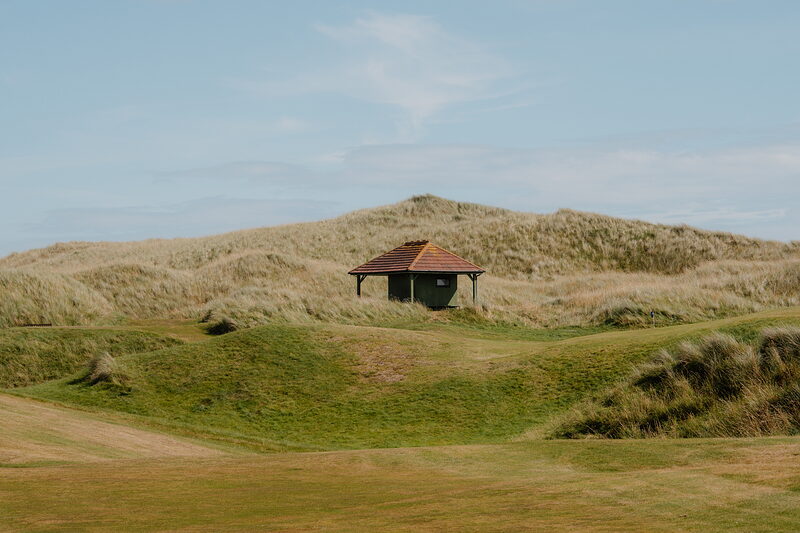 A dark timber shelter set into dunes beside a fairway, gorse bushes scattered across the mounds