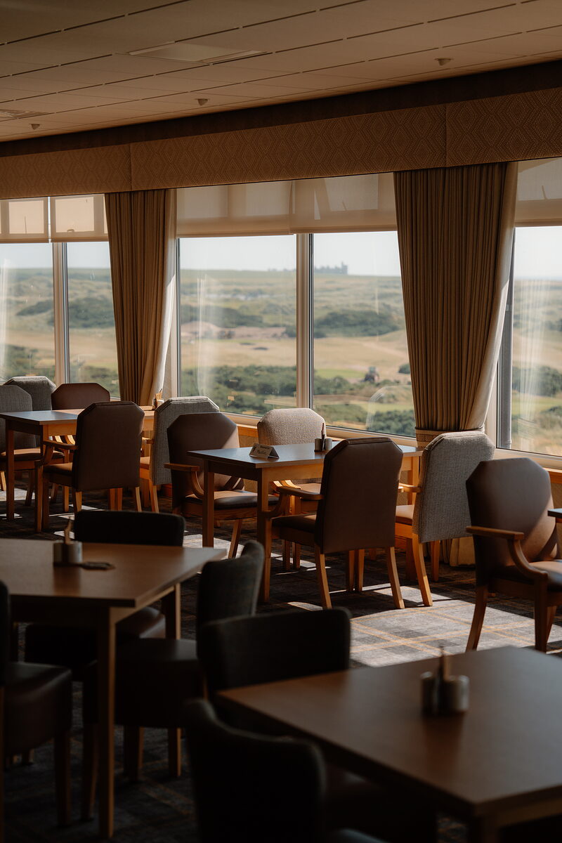 A dining room with upholstered chairs and tables set beside tall curtained windows overlooking the dunes