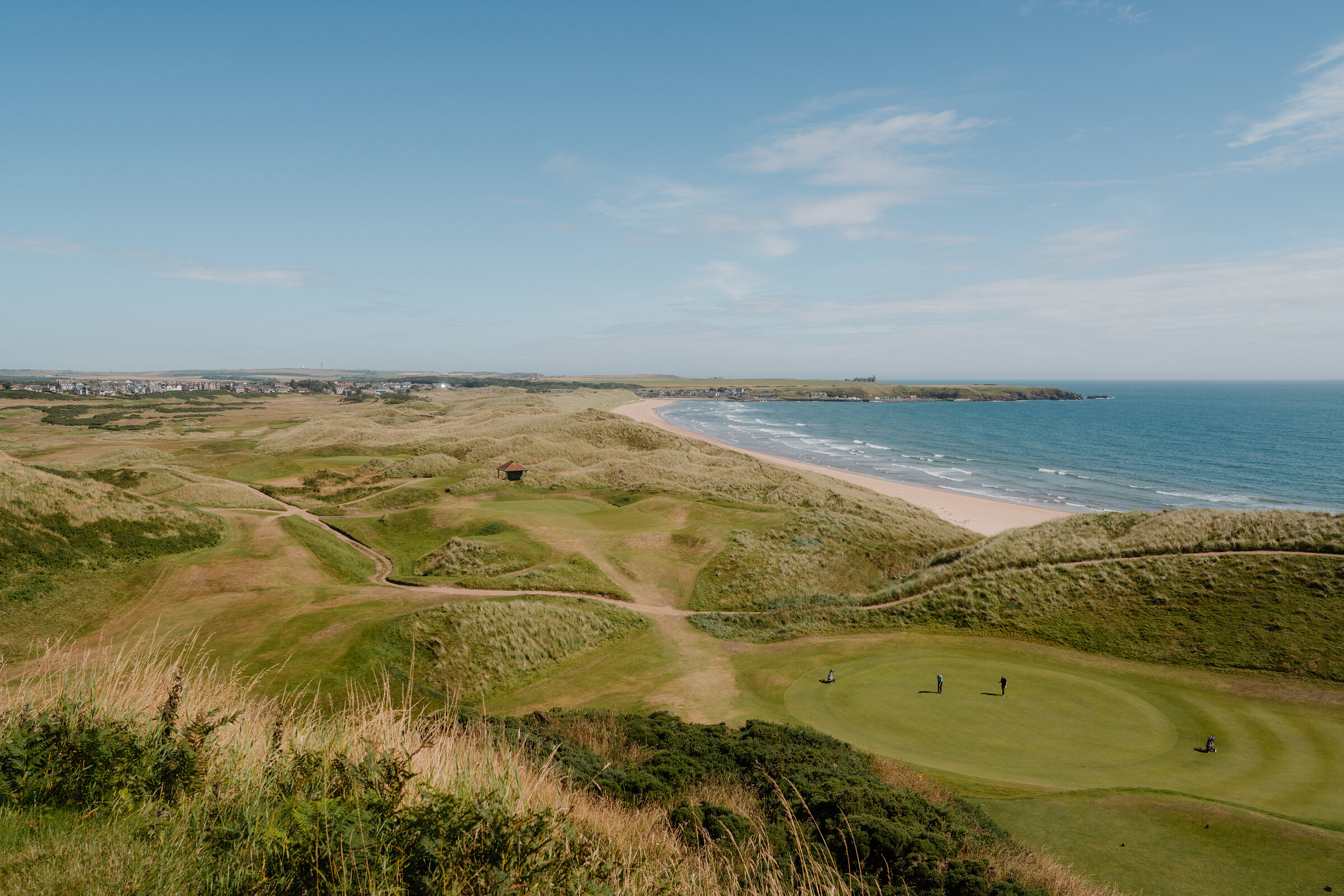 Golfers on a green overlooking a sandy beach and coastline, a town visible in the distance