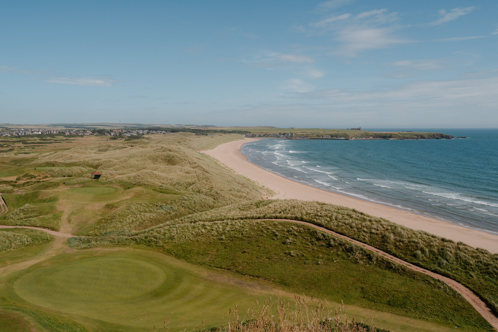 A green set among dunes overlooking a curving sandy beach and a coastal town