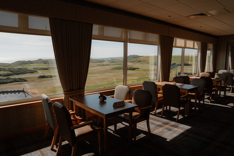 A tartan-panelled clubhouse room with round tables and chairs set beneath curtained windows overlooking the course