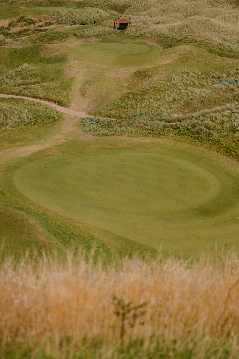 A green marked by a yellow flag set among dunes, a timber shelter visible on the rise beyond