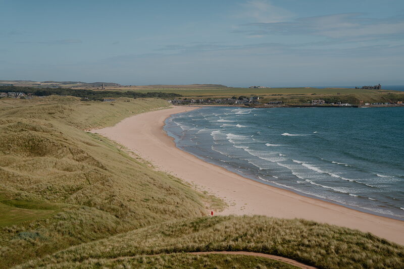 A curving sandy beach beneath grassy dunes, distant figures scattered along the shore