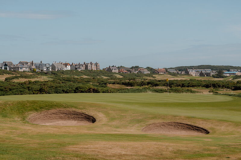 Twin bunkers set into a fairway, a row of sandstone buildings rising along the horizon