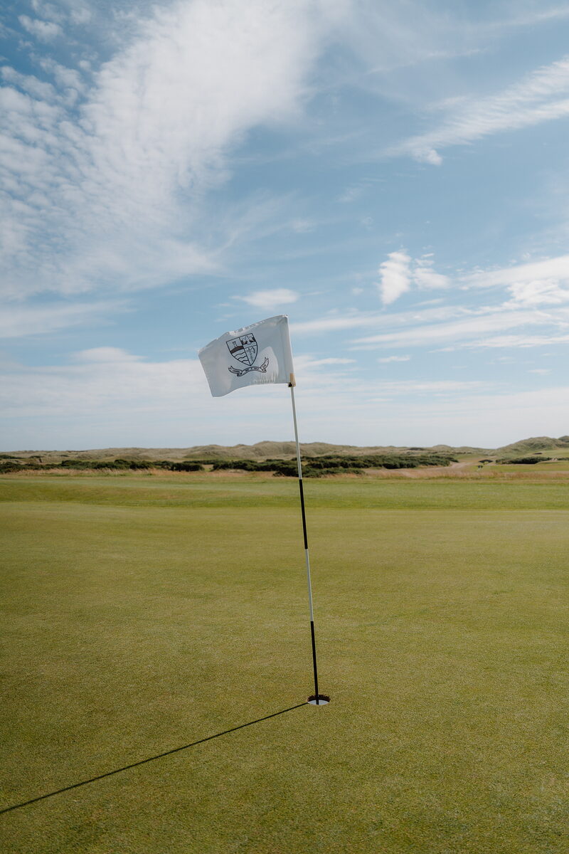 A white flag bearing a crest fluttering above a fairway bordered by dune grass