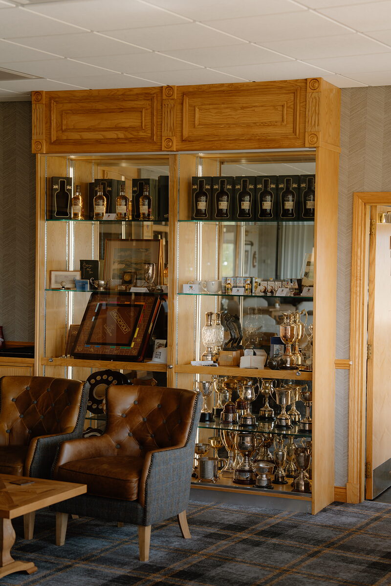 A glass display cabinet of whisky bottles and trophies beside leather armchairs