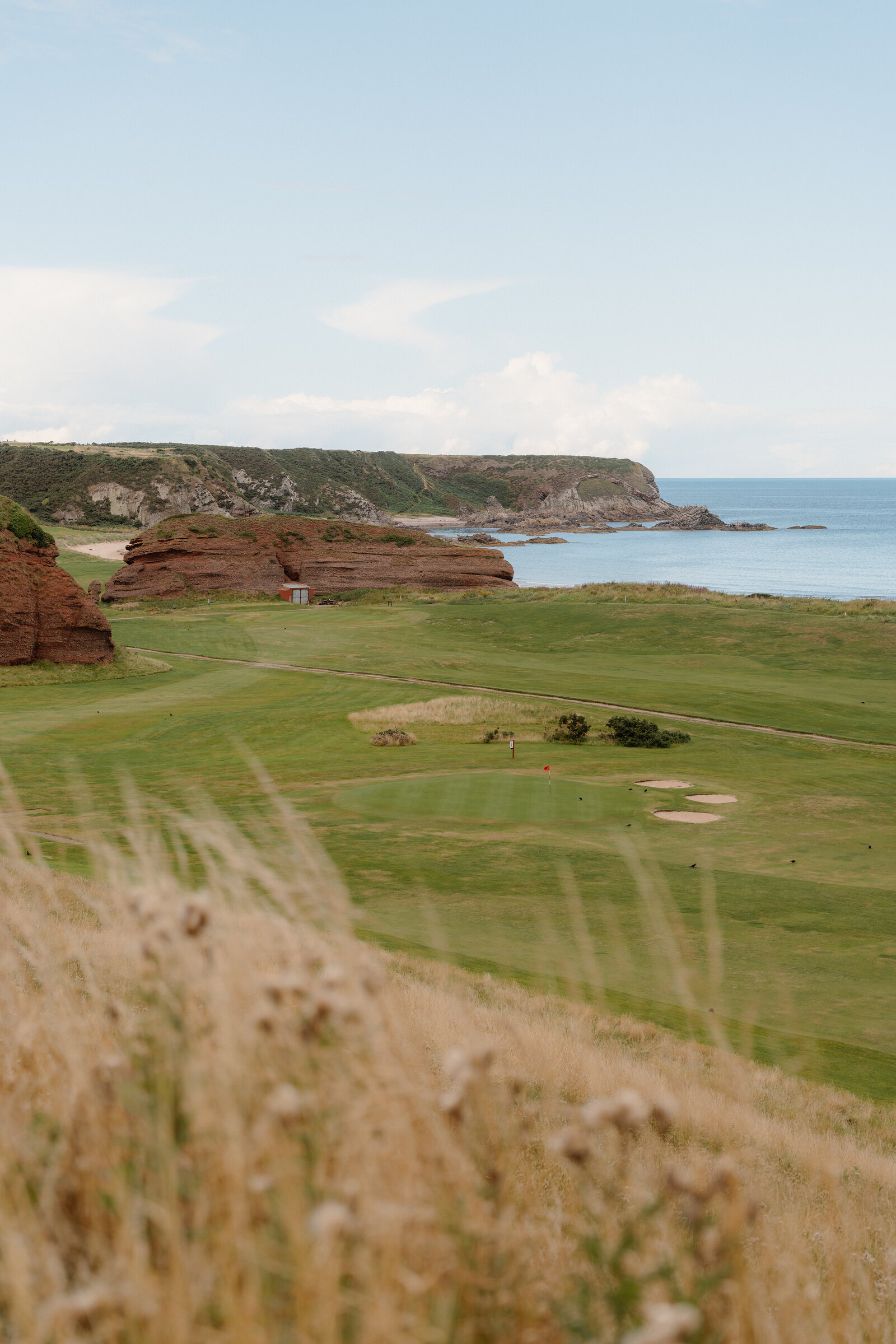 A coastal golf fairway leading towards red sandstone cliffs and the sea, dune grass in the foreground