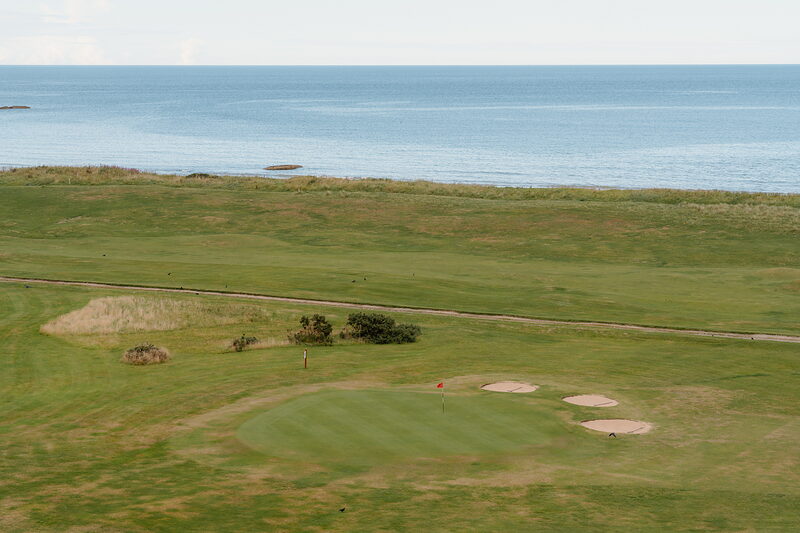 A golf green with a deep bunker overlooking the sea, hills fading into the distance