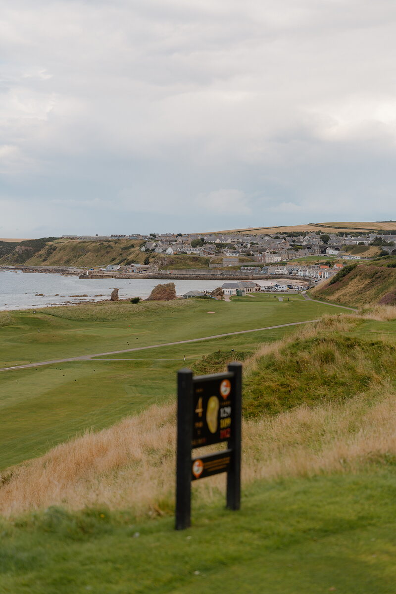 A hole marker sign overlooking a fairway and coastal village across the bay