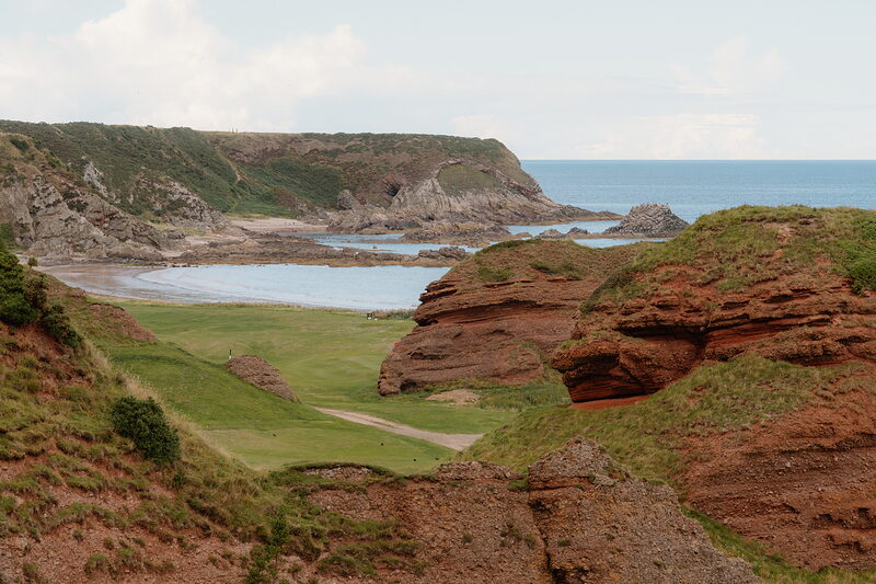 A fairway running between red sandstone cliffs down to a rocky cove and the sea