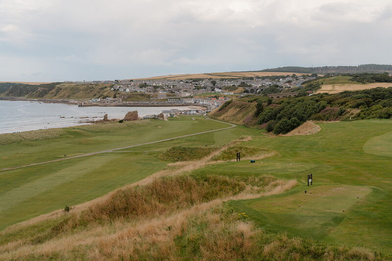 Golfers walking a fairway above a coastal town, a ruined tower standing on the shore below