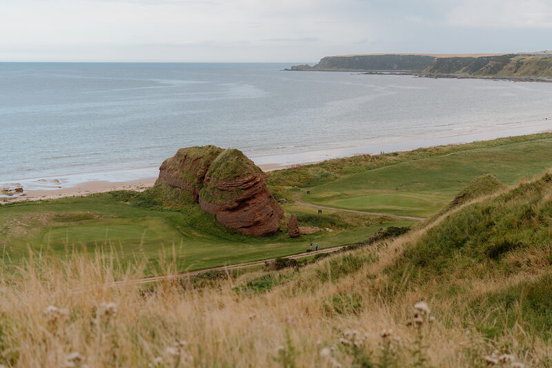 A red sandstone sea stack rising from a fairway beside a sandy beach and the sea