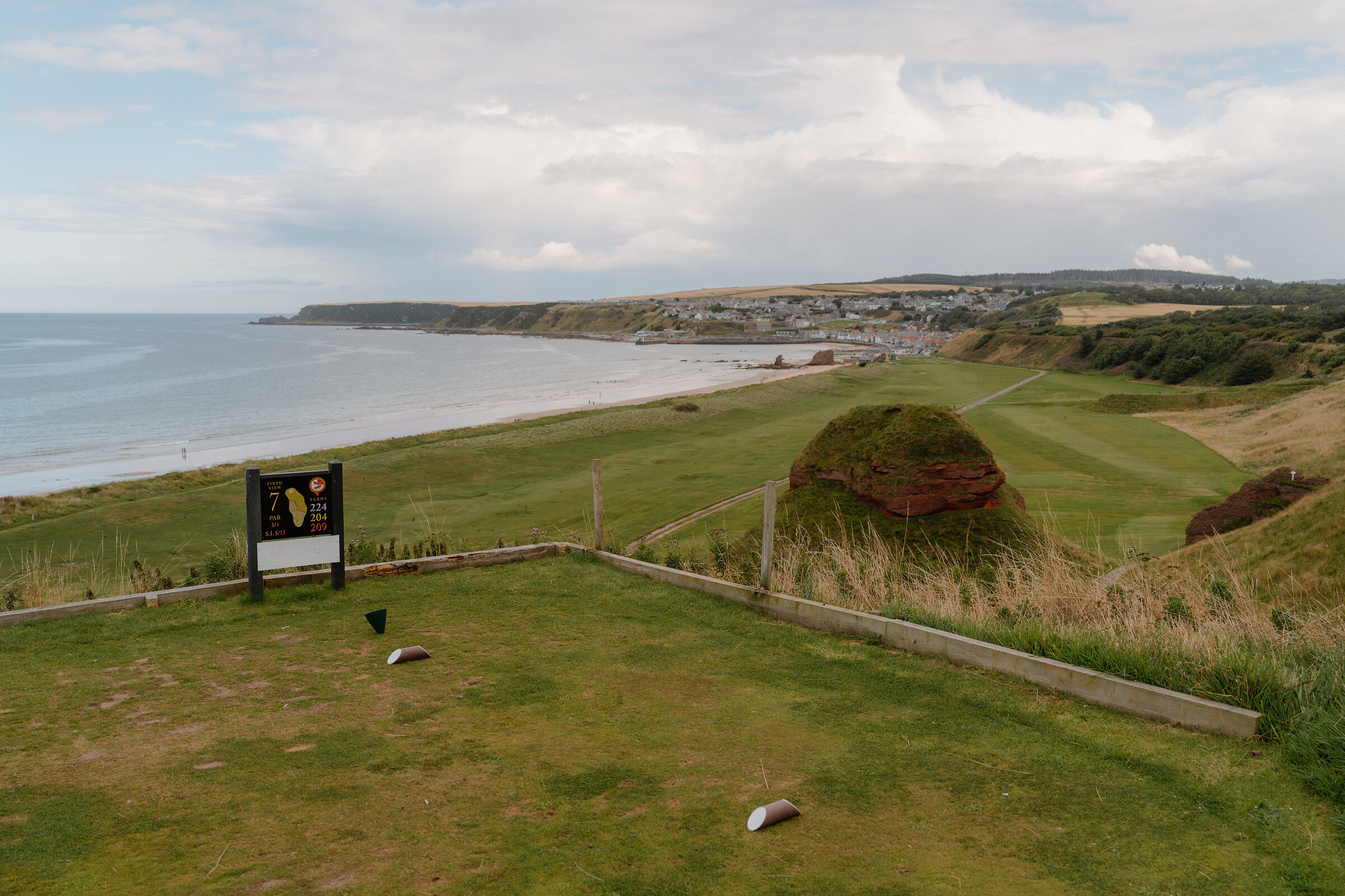 A tee marker sign overlooking a fairway and coastal town along the shore