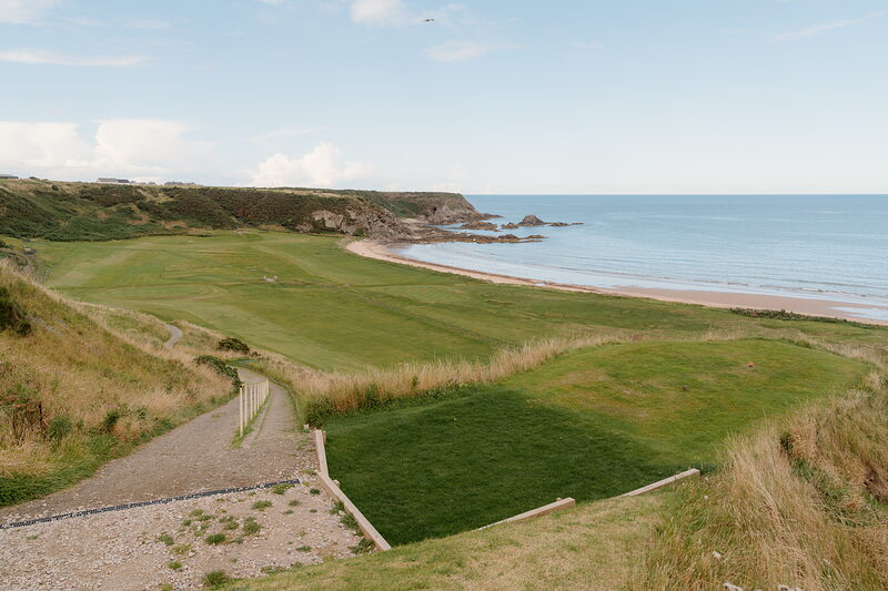 A gravel path leading down to a fairway bordered by dune grass, red cliffs and the sea beyond