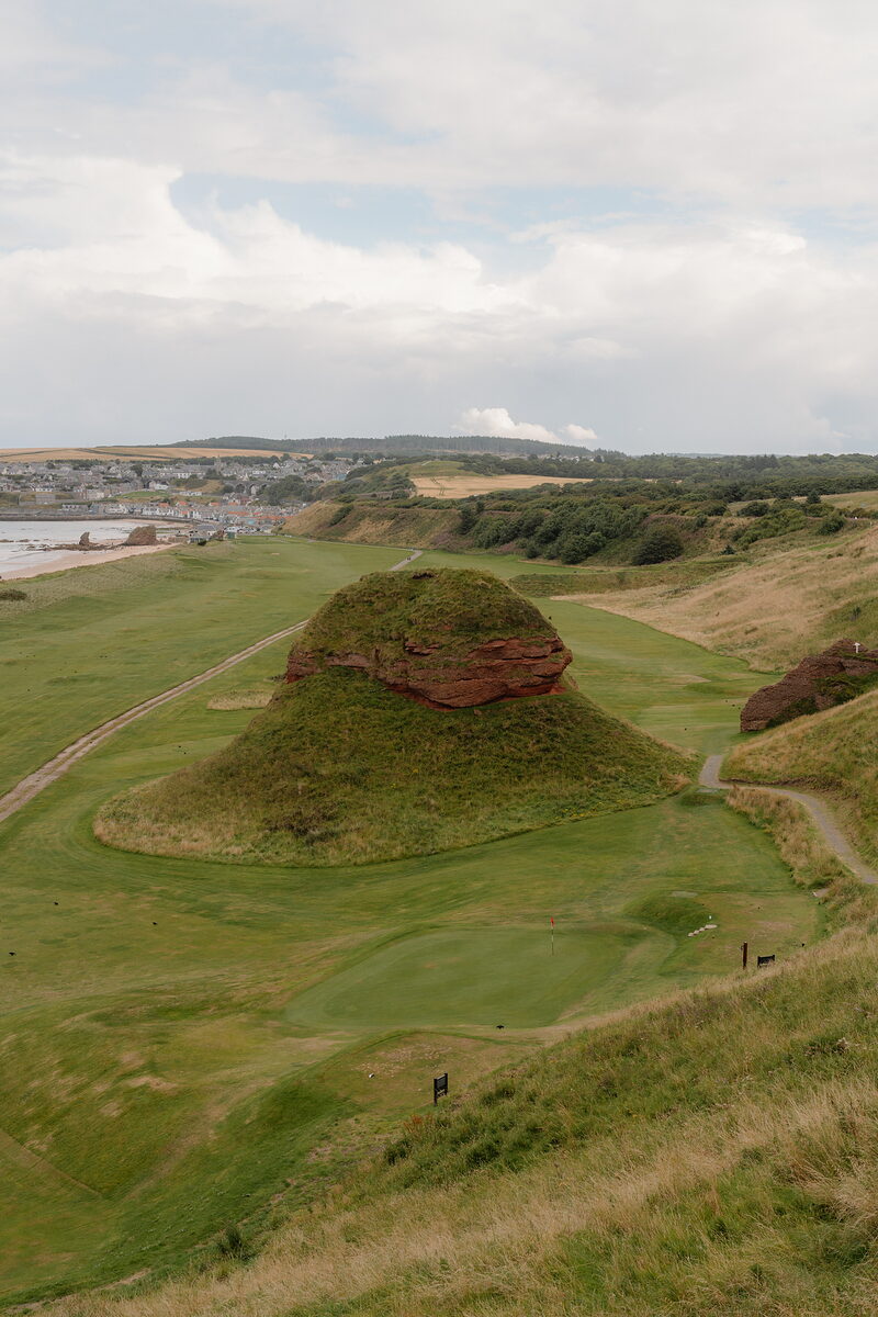 A red sandstone mound rising from a fairway, a coastal town and hills visible beyond