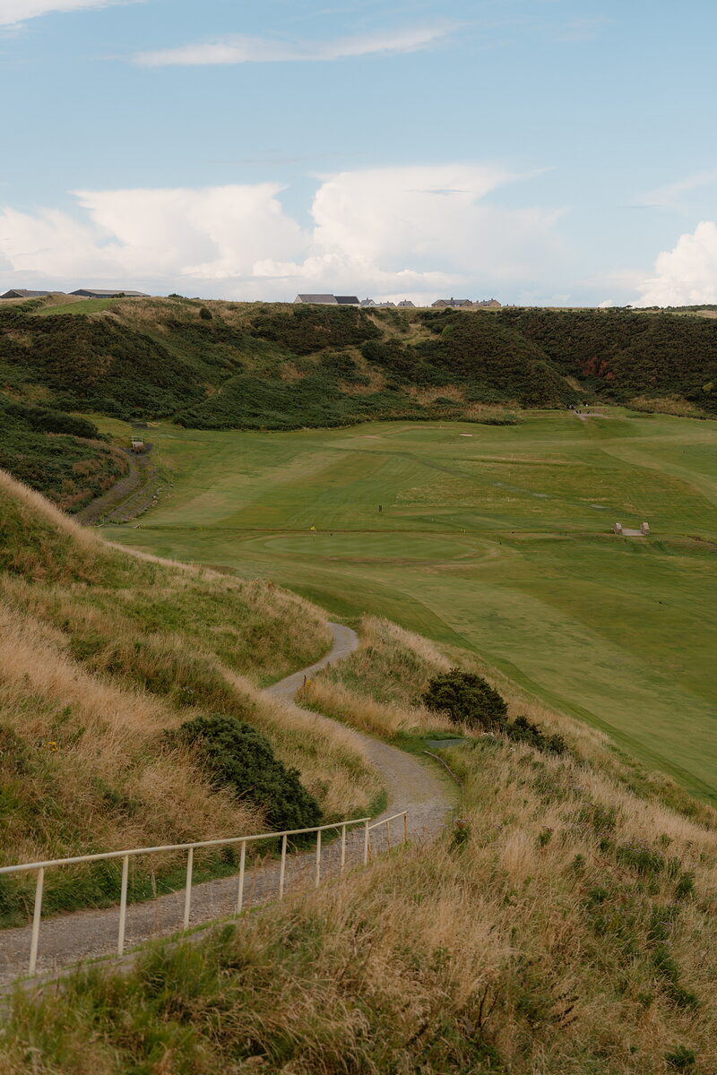 A gravel path winding along a fence beside a fairway set in a grassy hollow, buildings visible on the ridge above
