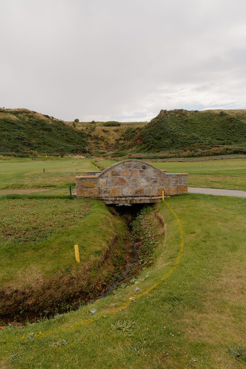 A stone bridge crossing a burn between two fairways, marker posts lining its banks