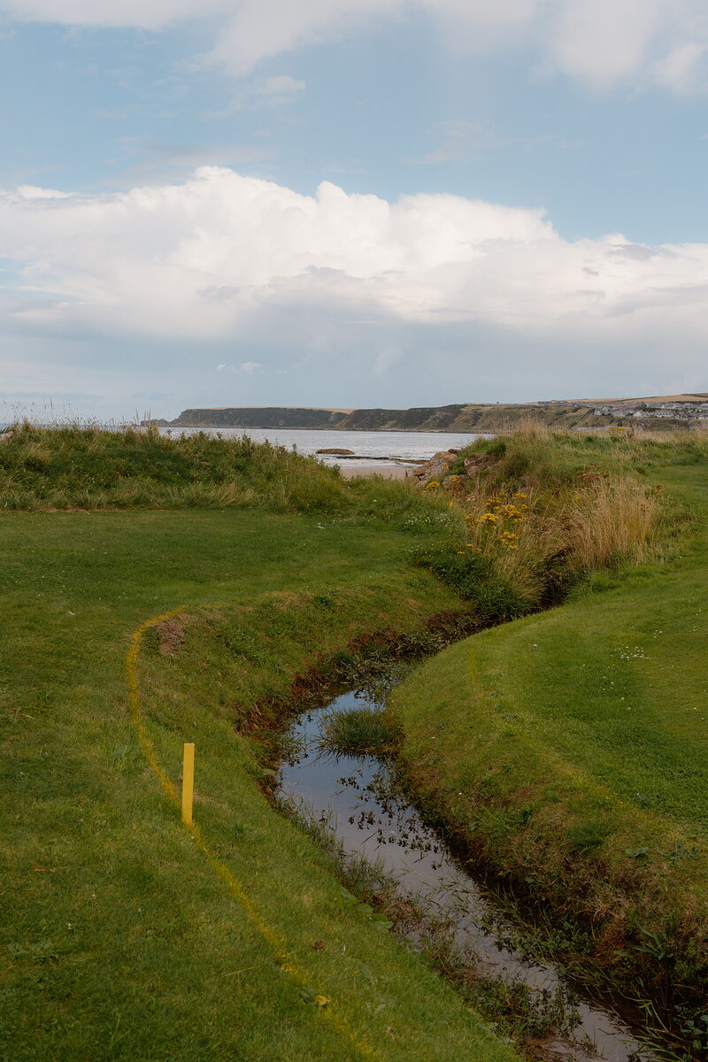 A stream curving through a fairway towards a bay bordered by cliffs and a distant town