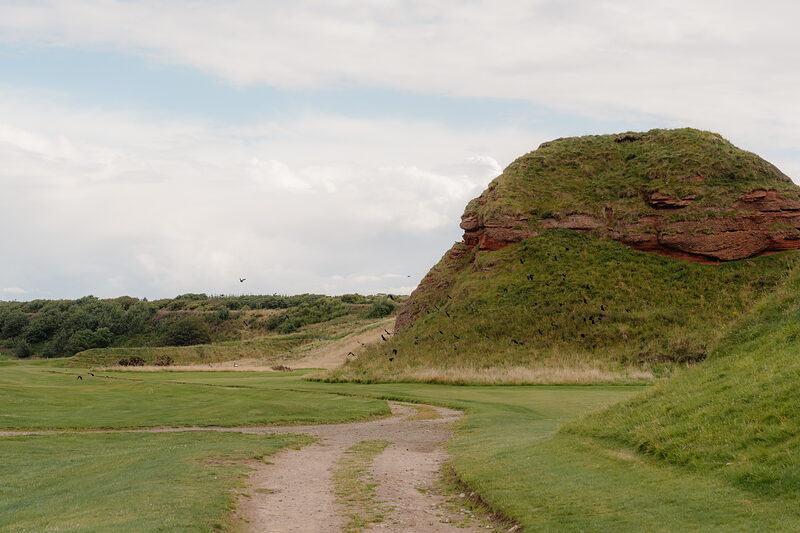 A gravel path curving past a grass-topped red sandstone outcrop beside the fairway