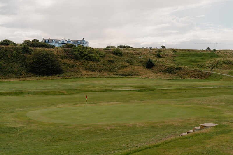 A pale blue clubhouse standing on a rise above a green marked by a red flag