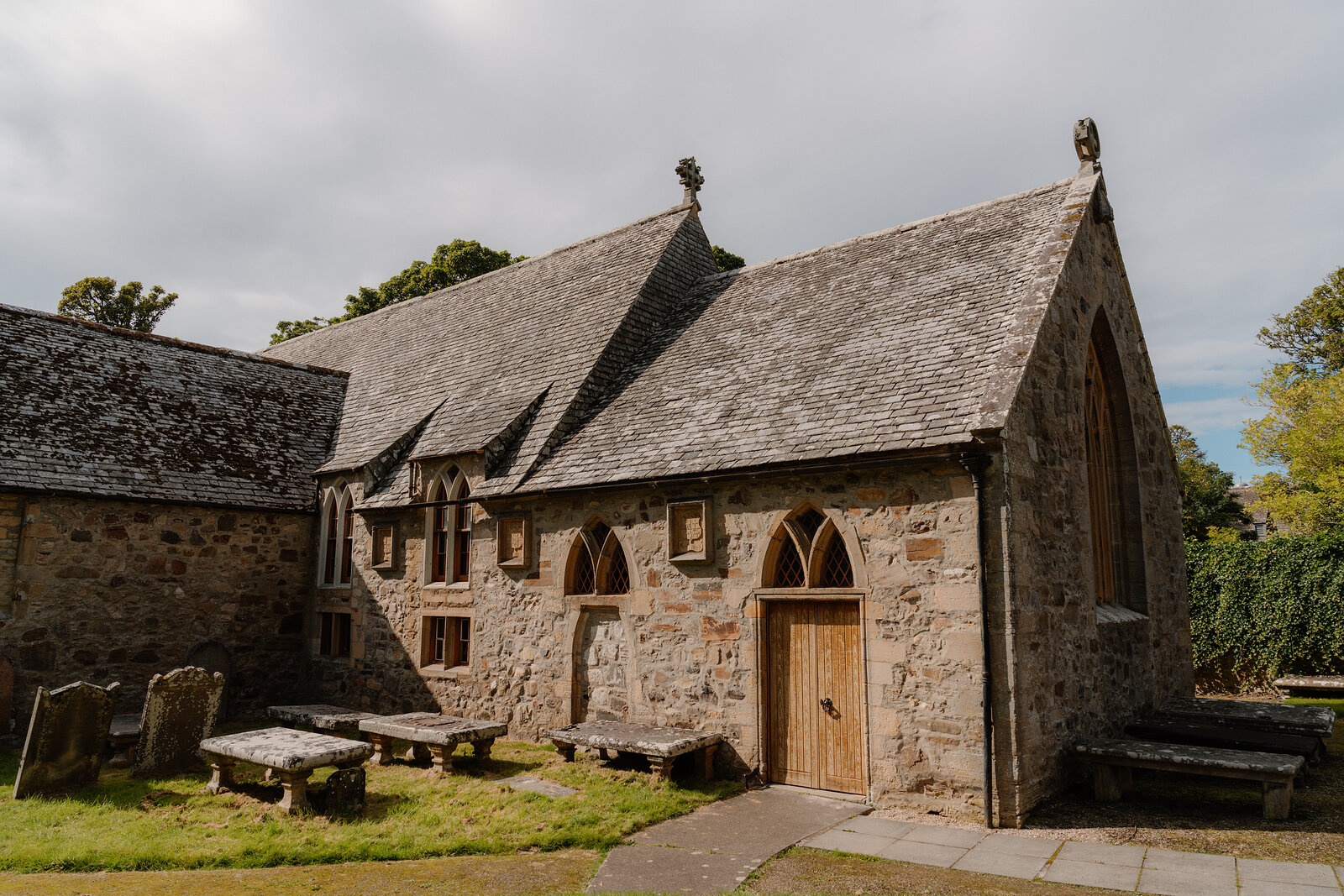 A stone church with arched windows and finials on its gable, gravestones set along the churchyard wall