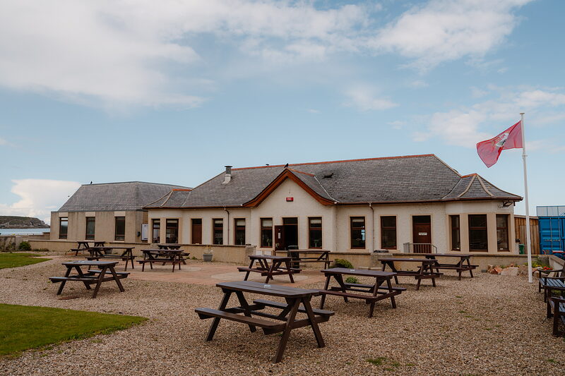 A pale clubhouse with a red-tiled porch, picnic tables set on the gravel forecourt beside a pink flag